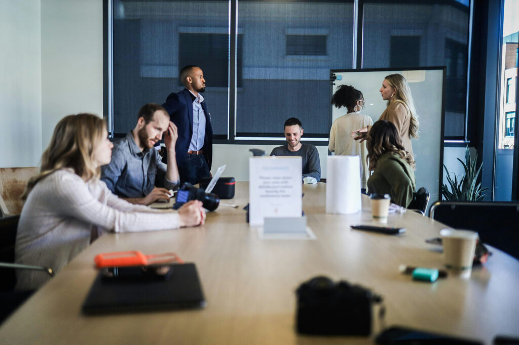 Group of coworkers gathered around a conference table, some seated and talking, others standing near a whiteboard.