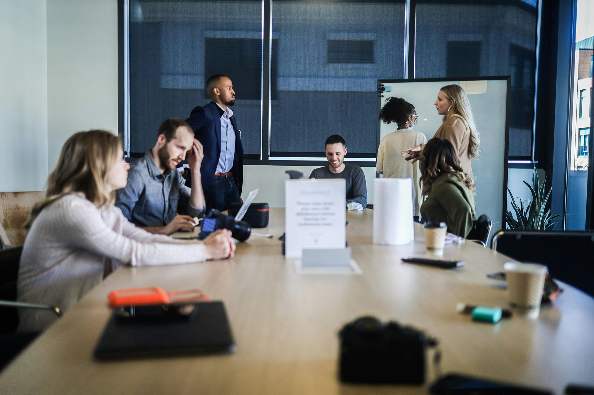 Group of coworkers gathered around a conference table, some seated and talking, others standing near a whiteboard.