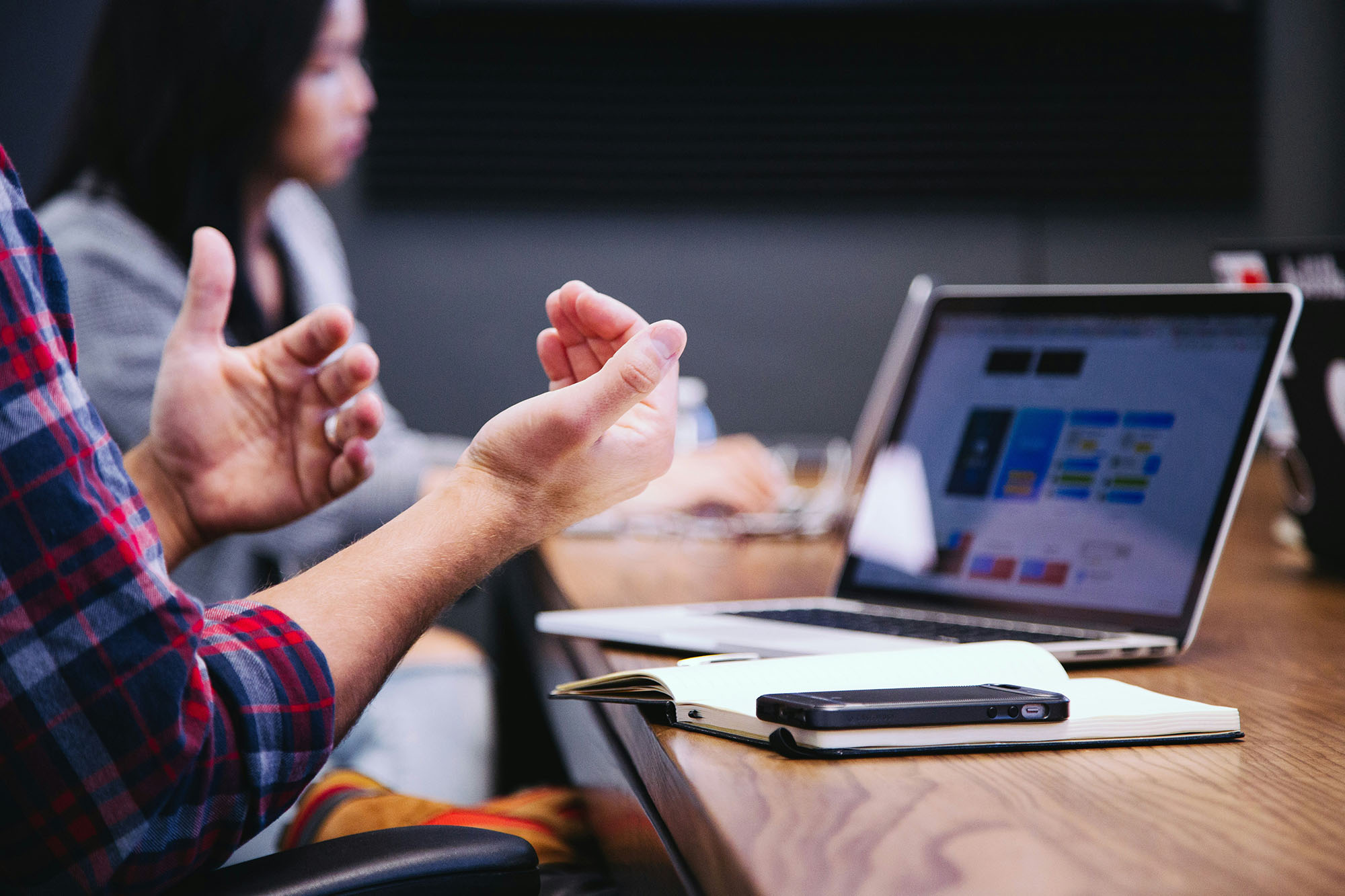 Person gesturing while talking in a meeting, with a laptop and notebook on the table.