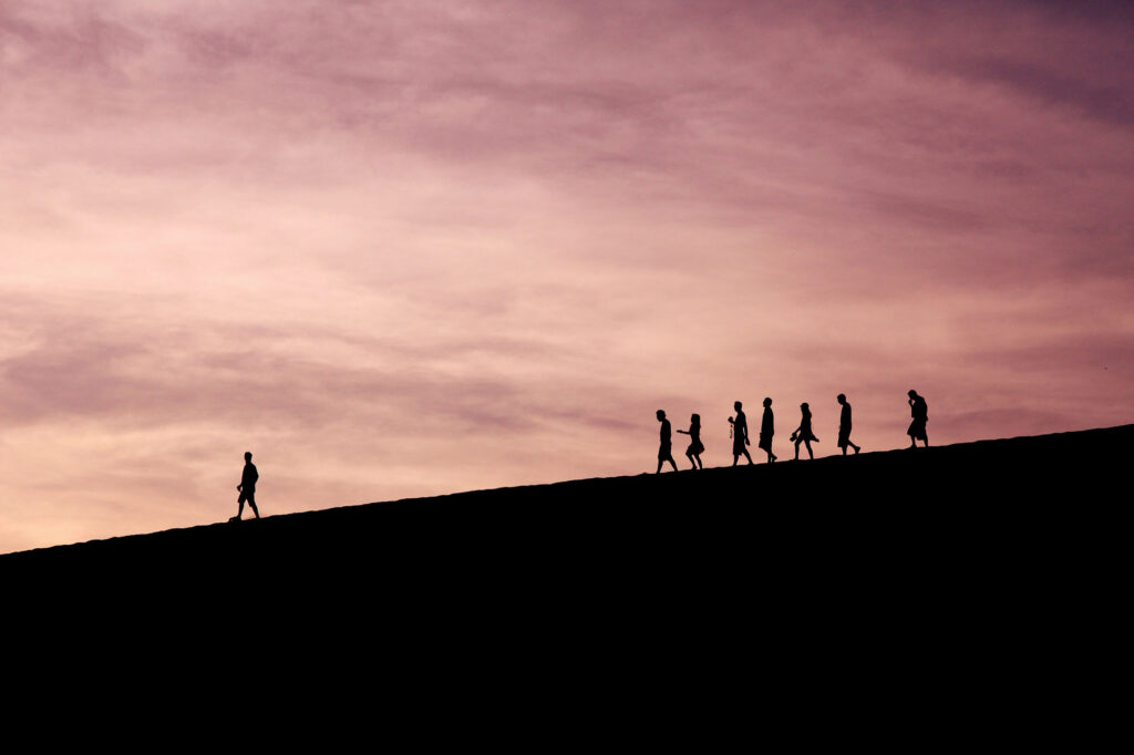 Silhouettes of people walking up a steep dune against a soft pink and purple sunset sky